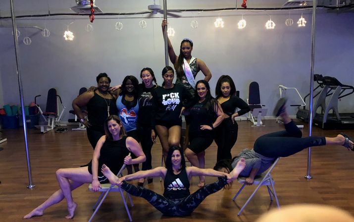Diverse group of women posing in a pole fitness studio around a vertical pole and folding chairs, wearing athletic dancewear on a wood floor with string lights — energetic group fitness vibe