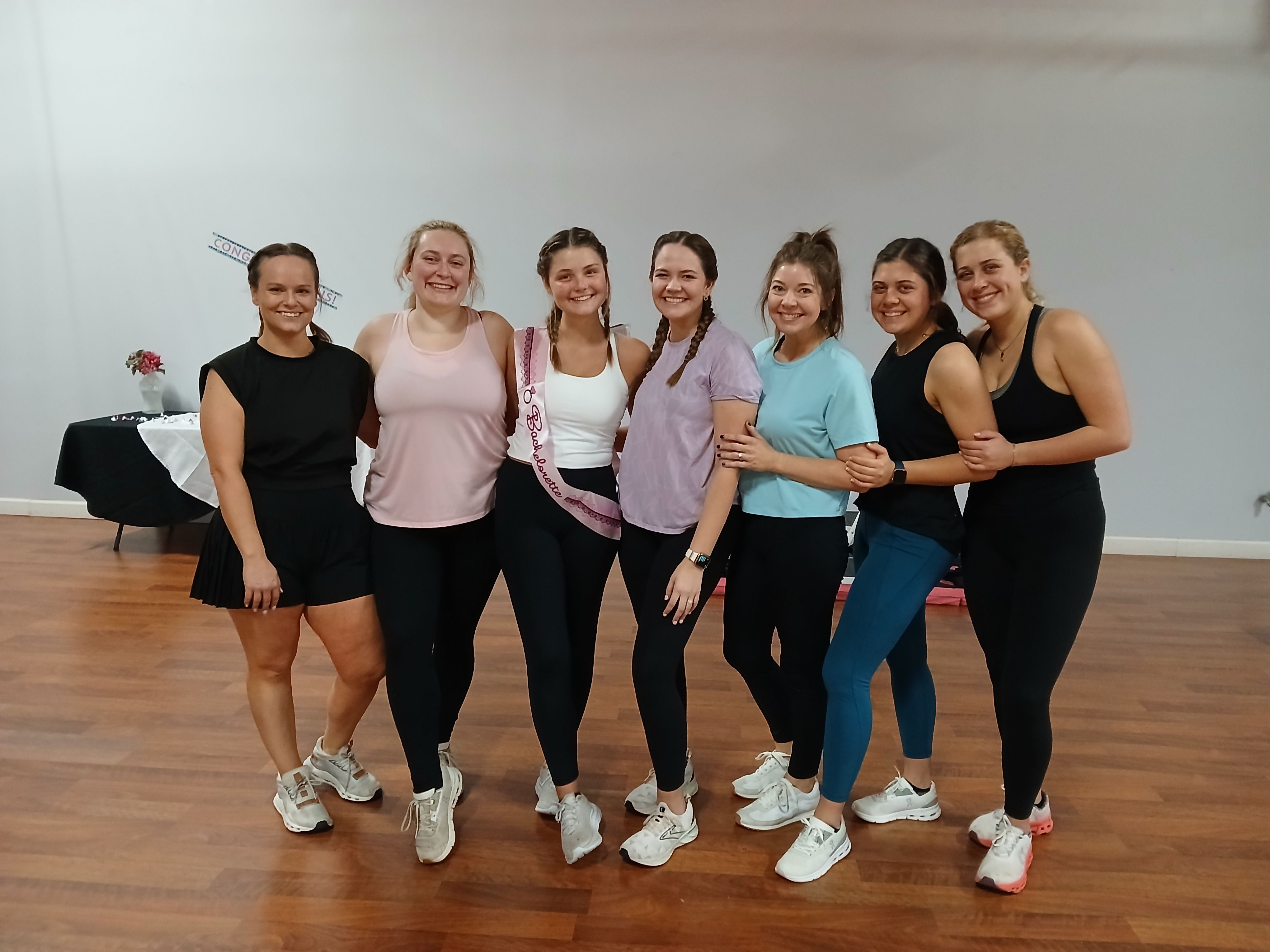 Seven friends in workout clothes smiling and posing in a bright dance studio with wood floors, one wearing a pink bachelorette sash.