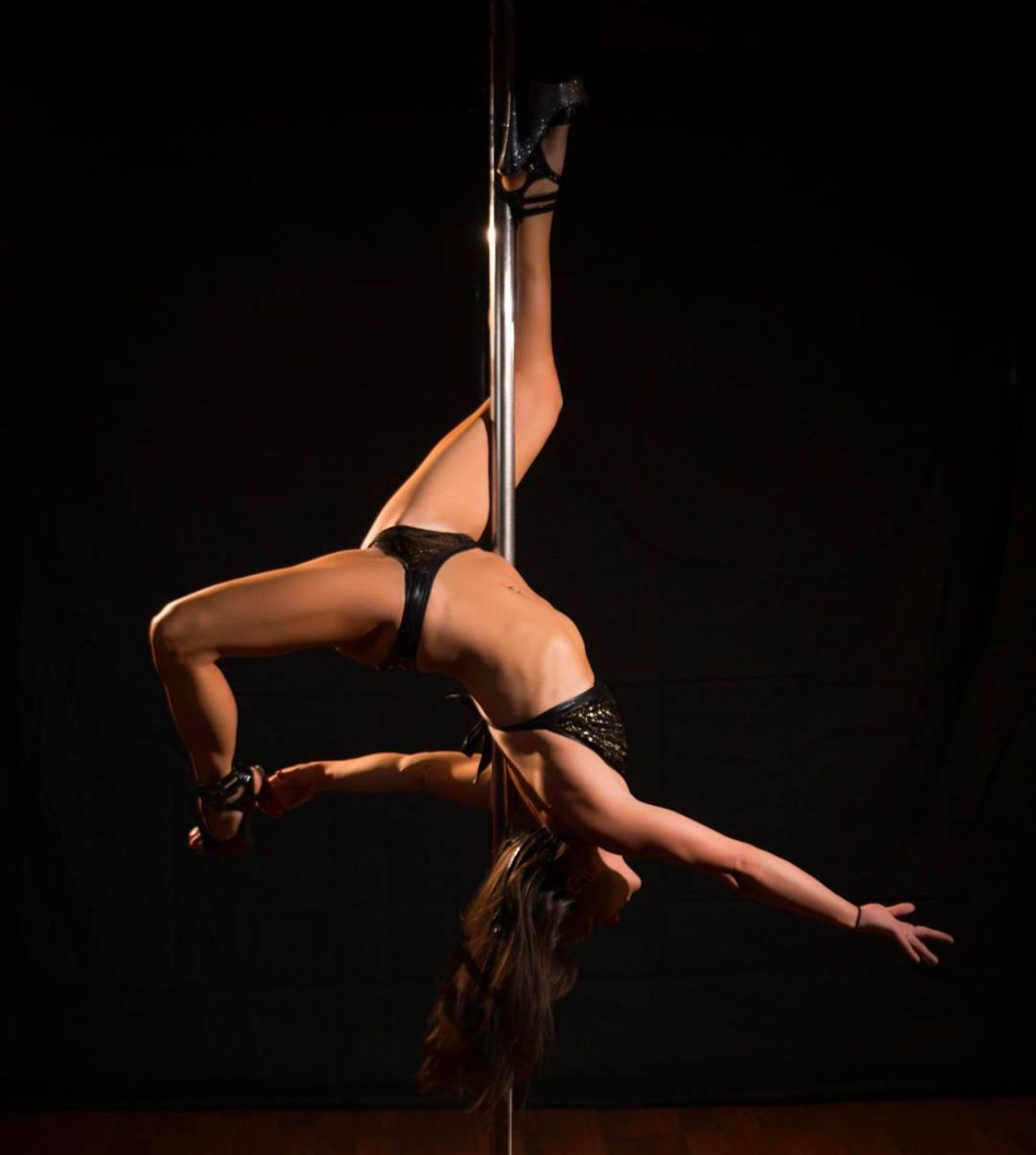 Graceful pole dancer in black costume and heels performing an inverted split on a vertical pole against a dark studio backdrop