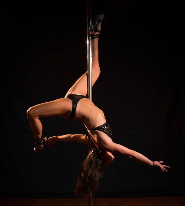 Graceful pole dancer in black costume and heels performing an inverted split on a vertical pole against a dark studio backdrop