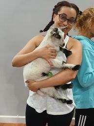 Smiling person with glasses and braids holding a white-and-black baby goat indoors — cozy petting-event moment