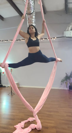 Midair aerial silks performer in a full straddle split on pink fabric inside an indoor aerial yoga studio — black sports bra, navy leggings, wood floor and string lights.