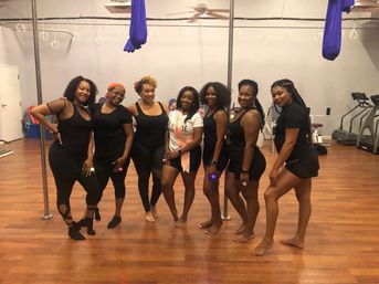 Seven women in activewear smiling and posing in a pole and aerial fitness studio with purple silks, metal poles and hardwood floor; one wears a sash for a group pole/dance workout.