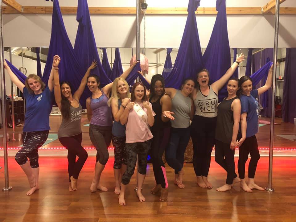 Ten women posing barefoot in an aerial yoga studio with purple hammocks and poles, smiling and celebrating on a wooden floor — one wearing a sash.