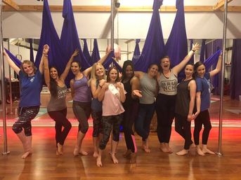 Ten women posing barefoot in an aerial yoga studio with purple hammocks and poles, smiling and celebrating on a wooden floor — one wearing a sash.