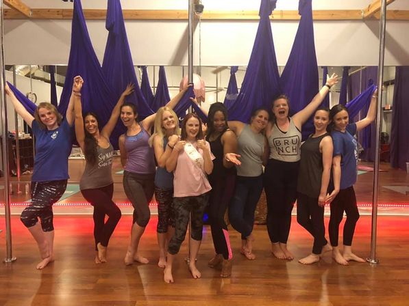 Ten women posing barefoot in an aerial yoga studio with purple hammocks and poles, smiling and celebrating on a wooden floor — one wearing a sash.