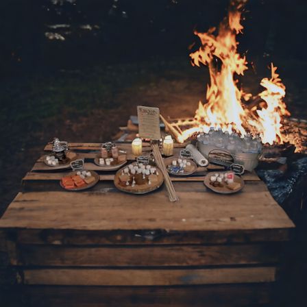Nighttime outdoor campfire dessert station on a rustic wooden table with roaring flames, plates of marshmallows and s'mores fixings, skewers, candles and chilled drinks.
