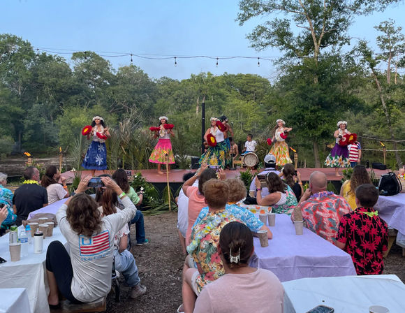 Festive outdoor luau with hula dancers in colorful skirts and leis performing on a riverside wooden stage with live drummers while an audience at picnic tables watches and takes photos under string lights in a wooded setting