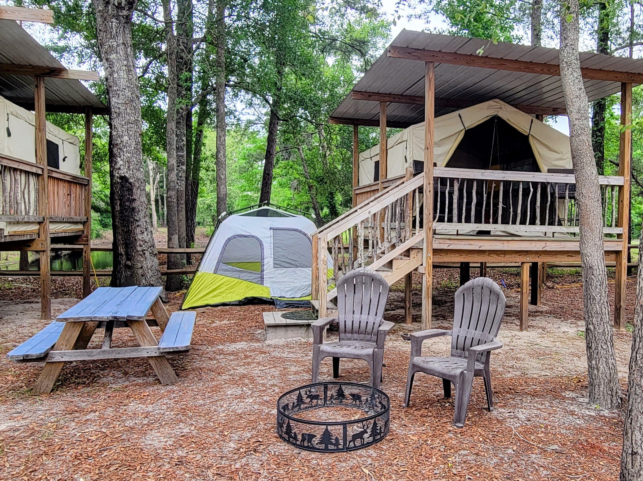 Cozy wooded campground scene with a raised canvas glamping tent on a wooden platform, a small ground tent, blue picnic table, two plastic chairs and a decorative metal fire pit