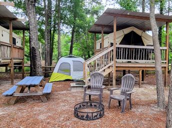 Cozy wooded campground scene with a raised canvas glamping tent on a wooden platform, a small ground tent, blue picnic table, two plastic chairs and a decorative metal fire pit