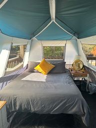 Glamping tent interior: queen bed with gray linens and a bright yellow pillow, wooden bedside table with lamp and yellow fan, large mesh windows overlooking a wooded deck with autumn foliage