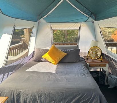 Glamping tent interior: queen bed with gray linens and a bright yellow pillow, wooden bedside table with lamp and yellow fan, large mesh windows overlooking a wooded deck with autumn foliage