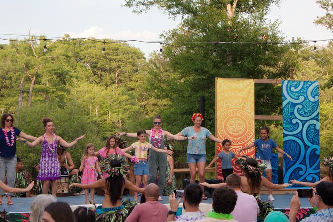 Outdoor luau-style dance on a wooden stage in a leafy park — hula performers and children wearing leis and colorful costumes, bright banners and string lights overhead at a family-friendly summer festival.