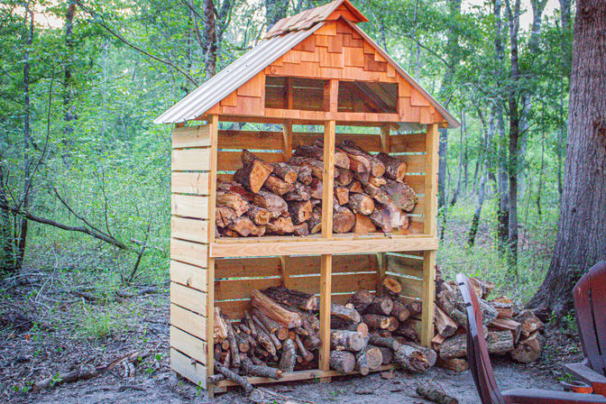 Rustic two-tier wooden woodshed with gabled roof neatly stacked with firewood at the edge of a green woodland backyard, ready for bonfire season.