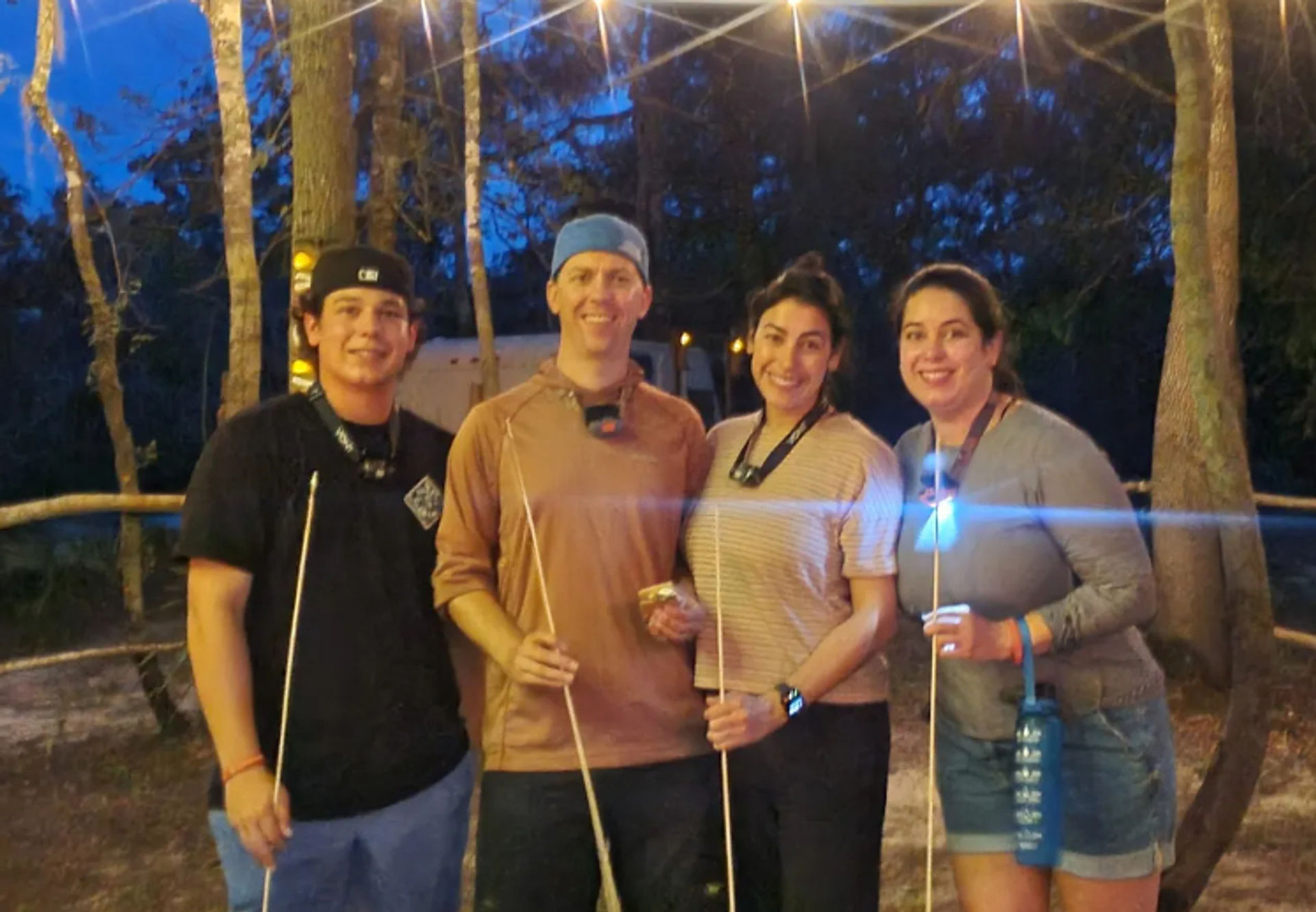 Four people smiling at a wooded campsite at dusk, holding marshmallow roasting sticks for s'mores under warm string lights during an evening bonfire gathering.