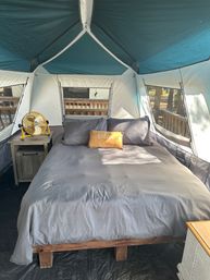 Sunlit glamping tent interior with a cozy queen bed in gray linens, yellow "relax" pillow, wooden nightstand with a yellow fan, and mesh windows overlooking a wooded deck.