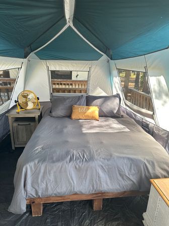 Sunlit glamping tent interior with a cozy queen bed in gray linens, yellow "relax" pillow, wooden nightstand with a yellow fan, and mesh windows overlooking a wooded deck.