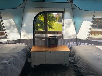 Cozy camping tent interior on a wooden deck with two beds flanking a storage chest topped by a portable heater, sunlight and green lawn visible through the window
