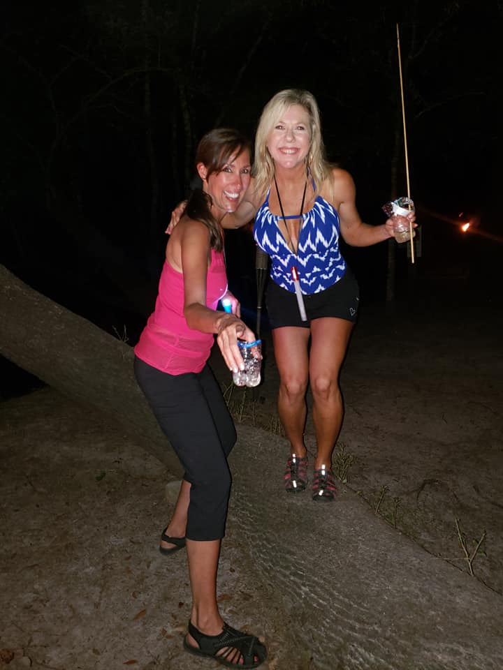 Two smiling women balancing on a fallen log on sandy ground at night, holding drinks and a lit sparkler stick in a dark outdoor setting