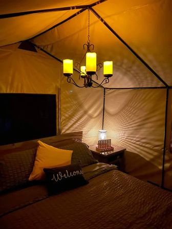 Cozy glamping tent interior at night with warm amber chandelier, bed with a welcome pillow and yellow accent cushion, and a bedside lamp casting rippled shadows on canvas walls.
