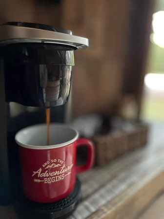 Close-up of a single-serve coffee maker pouring coffee into a red mug that reads 'And so the Adventure Begins' on a cozy home kitchen counter.