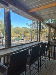 Covered wooden porch with high wicker bar chairs and a long counter overlooking picnic tables, colorful fabric banners, and a sunny wooded clearing under a clear blue sky.