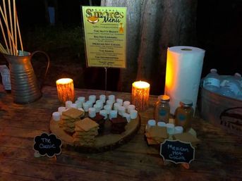 Outdoor candlelit s'mores station on a rustic wooden table at night with stacks of graham crackers, marshmallows, chocolate squares, jars of toppings, roasting sticks and a printed s'mores menu