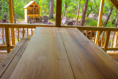 Rustic wooden table on a cabin porch overlooking a wooded backyard with Adirondack chairs around a fire pit and a stacked wood shed