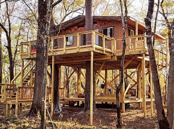 Rustic wooden treehouse cabin on tall stilts with wraparound deck and stairs tucked among sunlit trees in a cozy forest retreat.