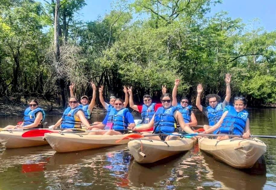 Group of kayakers in blue life jackets on a sunny, tree-lined river raising hands in celebration.