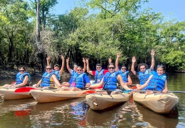 Group of kayakers in blue life jackets on a sunny, tree-lined river raising hands in celebration.