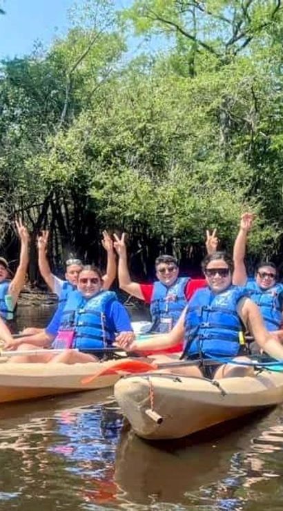 Group of kayakers in blue life jackets on a sunny, tree-lined river raising hands in celebration.