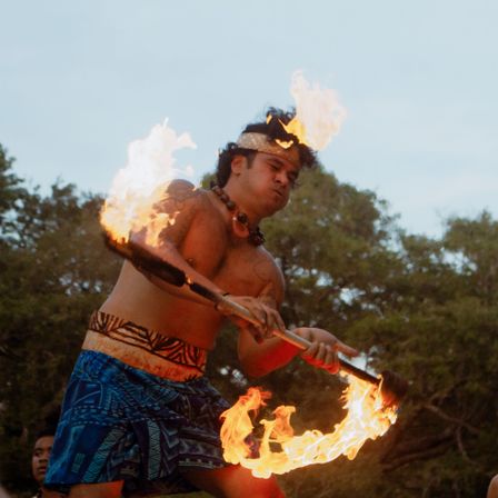 Island fire dancer spinning a flaming baton at dusk, shirtless performer in patterned sarong and headband creating dramatic fire trails against a tropical tree backdrop