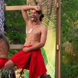 Shirtless dancer with tribal face paint sticking out his tongue, wearing a red wrap skirt and shell necklace during an energetic outdoor cultural dance on a green patterned stage backdrop.
