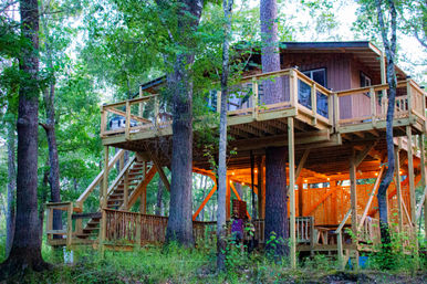 Cozy elevated wooden treehouse cabin on stilts with wraparound deck and stairs, warmly lit under-structure string lights, nestled among tall leafy trees in a lush forest at dusk.