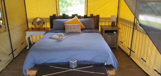 Cozy glamping tent interior with blue-dressed queen bed, yellow canvas walls, wooden nightstands with vintage fan and lamp, decorative pillows and a breakfast tray on the bed, and wood-look flooring.
