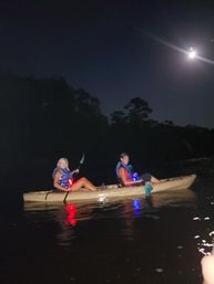 Two people paddling a tandem kayak on a moonlit lake at night, wearing life jackets with red and blue safety lights reflecting on the water.