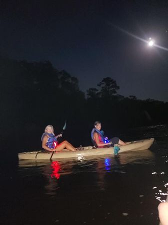 Two people paddling a tandem kayak on a moonlit lake at night, wearing life jackets with red and blue safety lights reflecting on the water.