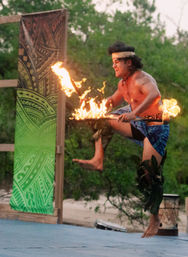 Shirtless Polynesian-style fire dancer leaping on an outdoor stage, twirling flaming torches with a green patterned backdrop and tropical trees