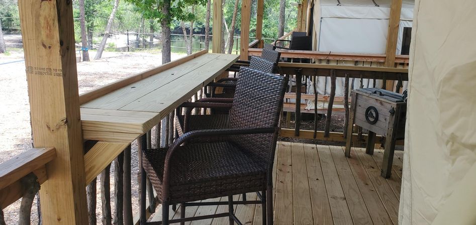 Glamping-style wooden deck with bar-height counter, woven wicker bar stools, rustic twig railing and canvas tent, overlooking trees and a small pond