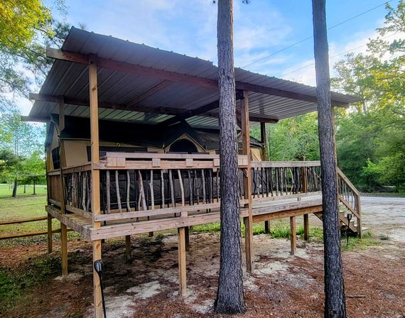 Raised rustic wooden deck with stick-railings and steps hosting a cozy canvas glamping tent under a corrugated metal roof, nestled among pine trees in a wooded campsite.