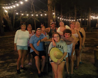 Group of friends and family posing at a wooded outdoor campground at night under string lights, gathered around a picnic table; several wear headlamps, one holds a straw hat and another wears a Texas sweatshirt.