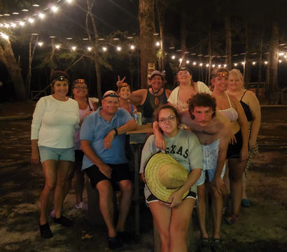Group of friends and family posing at a wooded outdoor campground at night under string lights, gathered around a picnic table; several wear headlamps, one holds a straw hat and another wears a Texas sweatshirt.
