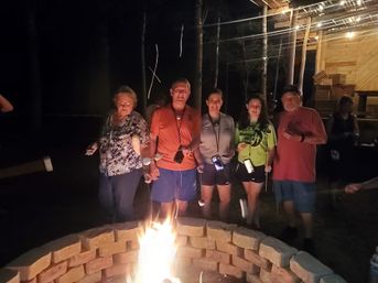 Five people gathered around a brick fire pit at night, roasting marshmallows on sticks near a wooden cabin lit with string lights, phones glowing.