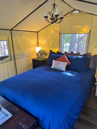 Cozy glamping tent bedroom with queen bed in blue bedding, rustic wood headboard, bedside table with lamp and metal fan, chandelier overhead, and window views of pine trees at a wooded campsite.