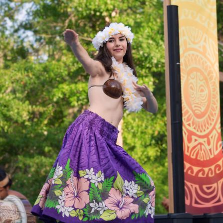 Hawaiian hula dancer in a purple hibiscus skirt, coconut-shell top and white flower lei performing on an outdoor stage with a tiki banner and lush green backdrop.
