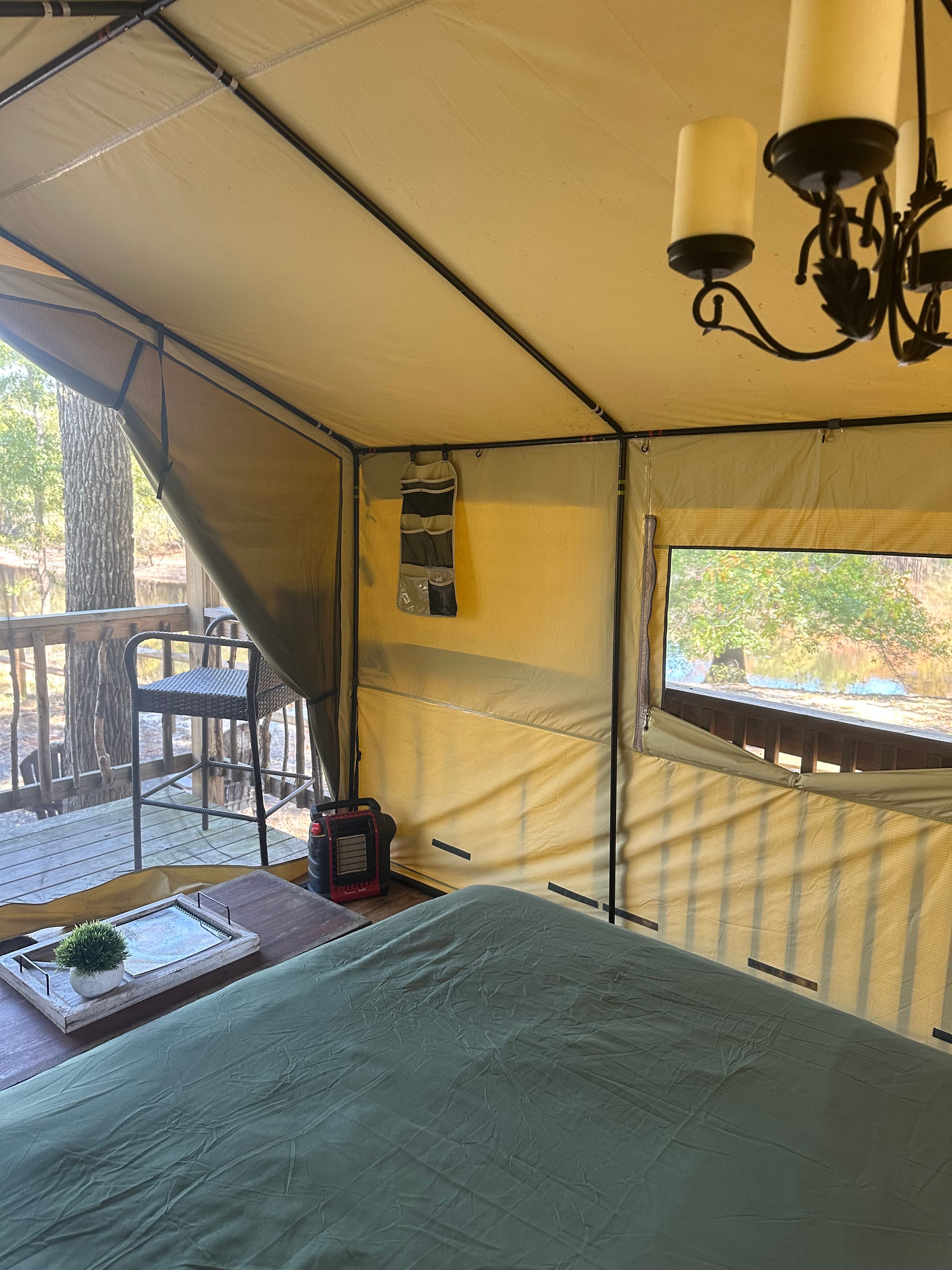 Glamping tent interior on a wooden deck: green-sheeted bed, wooden side table with tray and potted plant, portable heater, hanging organizer and chandelier, open mesh window with tree-lined pond view.