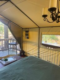 Glamping tent interior on a wooden deck: green-sheeted bed, wooden side table with tray and potted plant, portable heater, hanging organizer and chandelier, open mesh window with tree-lined pond view.