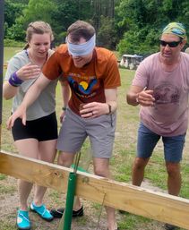 Blindfolded man guided by two teammates during a playful outdoor team-building challenge in a grassy park