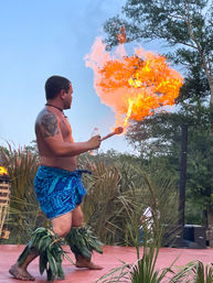 Polynesian fire dancer in blue sarong and leaf anklets spitting a large flame on an outdoor luau stage with palm foliage
